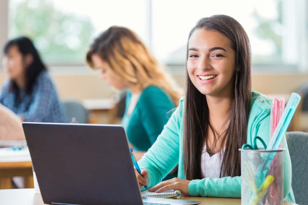Pretty Hispanic high school student works on project in graphic design class. She is looking at the camera and smilng as she takes notes. A laptop is open on the table and art supplies are also on the table. Students are working in the background.